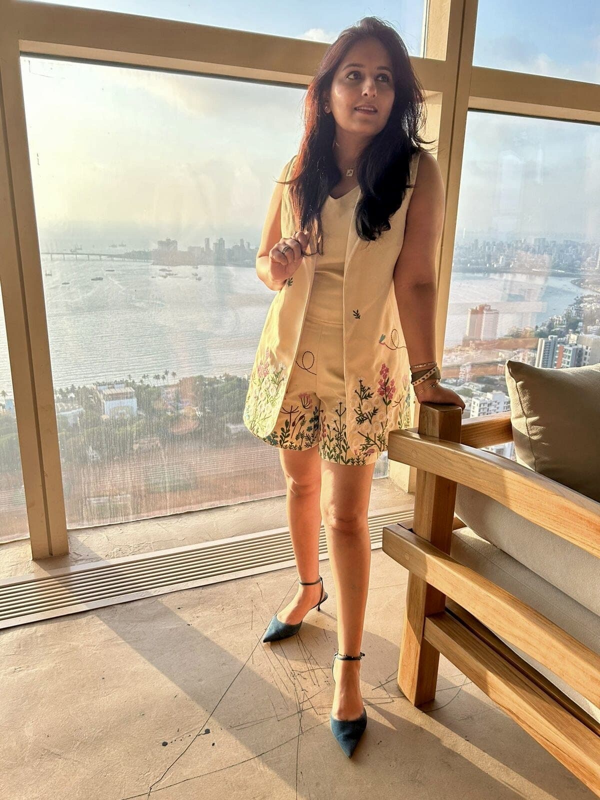Woman wearing the cream garden-embroidered set — long open sleeveless jacket over camisole and shorts — beside a tall sea-facing window.