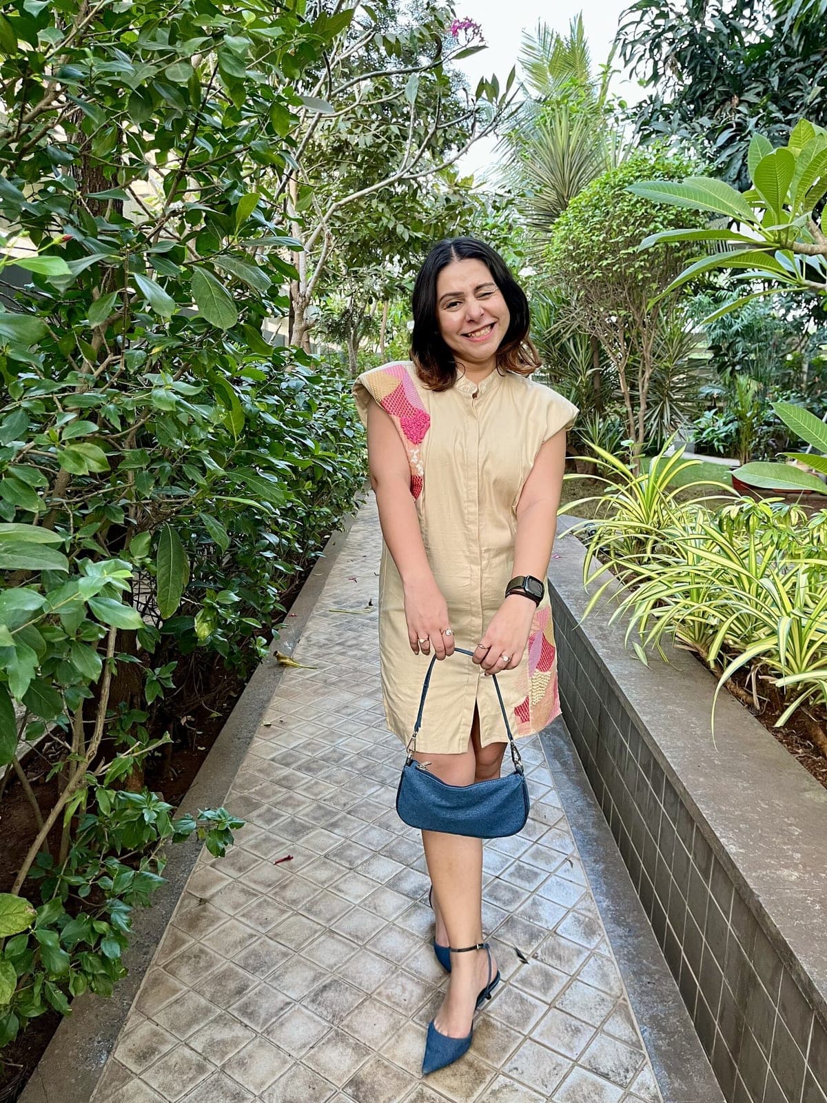 Sand-coloured sleeveless shirt dress with padded shoulders and pink patchwork embroidery down one shoulder, worn on a garden path.