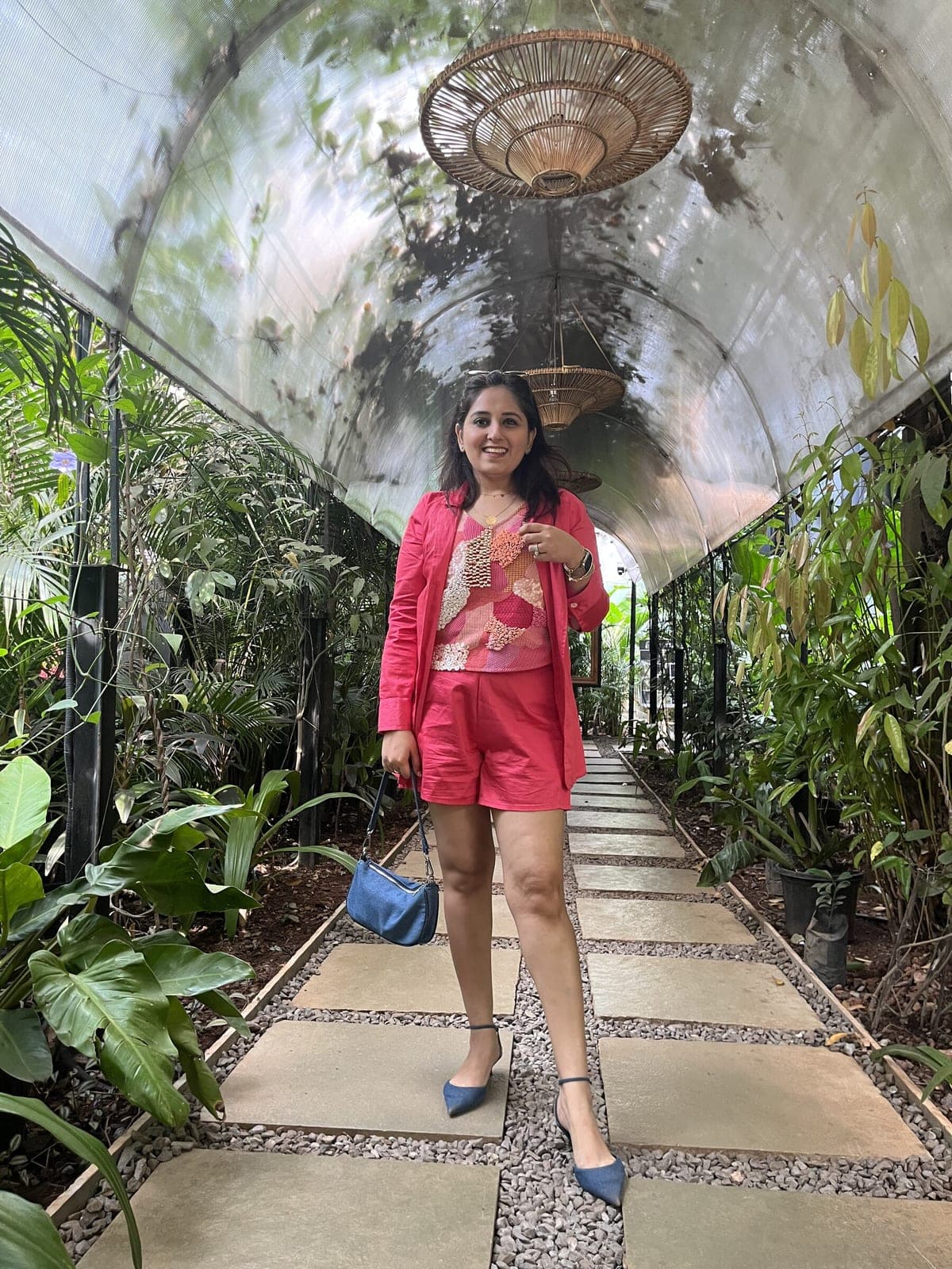 Woman standing in a greenhouse walkway in an open pink blazer over a beaded patchwork top and matching pink shorts.