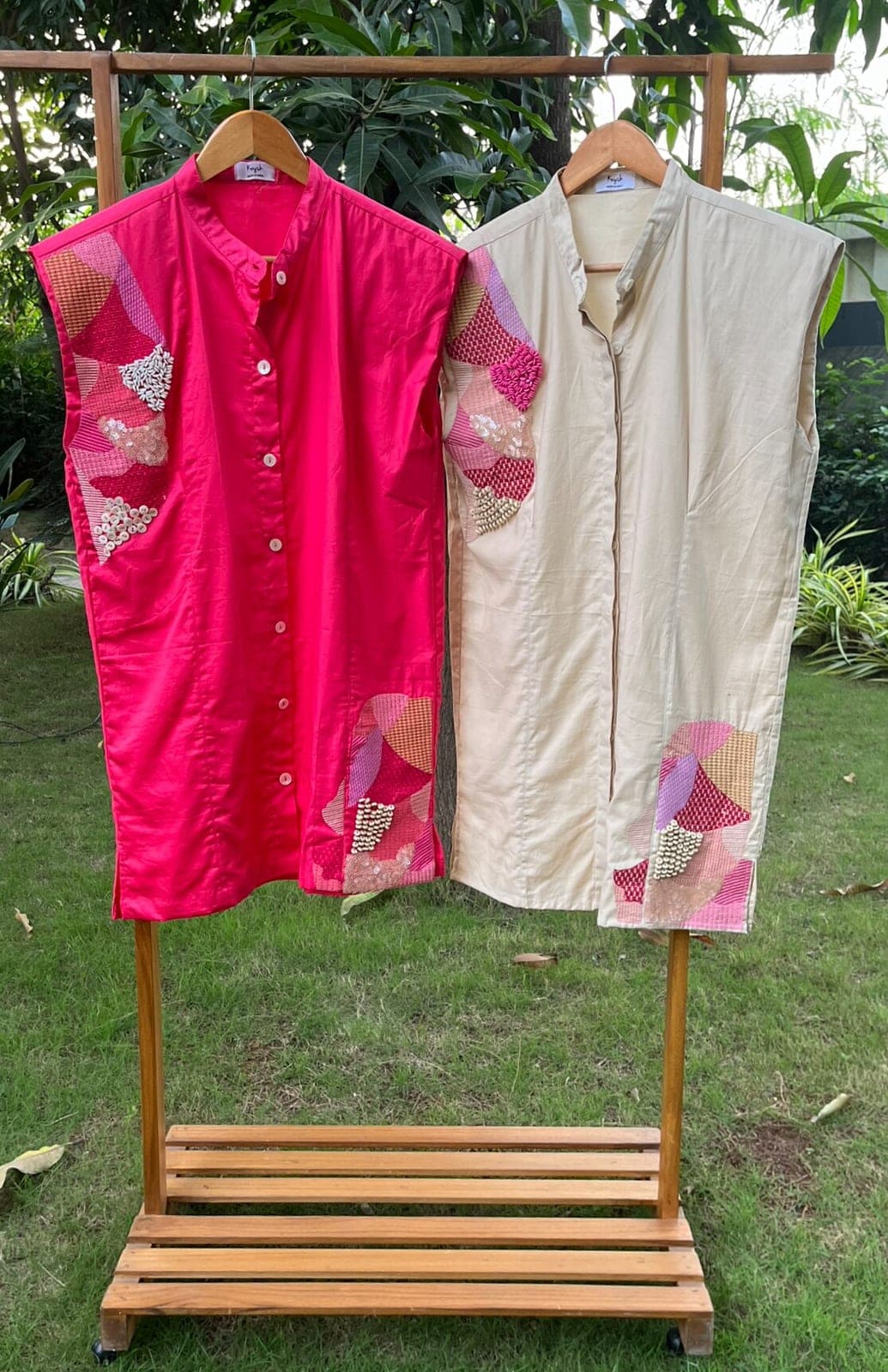 Two sleeveless button-front dresses on a wooden rack, one deep pink and one cream, both beaded with patchwork at shoulder and hem.