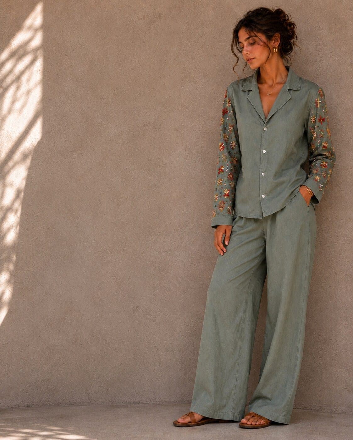 Standing against a beige wall, a model wears the embroidered shirt with matching wide-leg trousers in the same grey-green cotton.