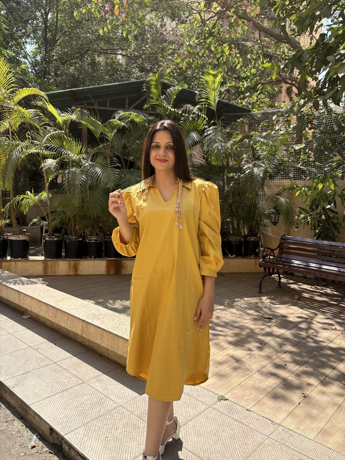 Woman in a paved courtyard wearing the mustard dress alone: collared split neck, beaded cord ties, gathered three-quarter sleeves, hem below the knee.