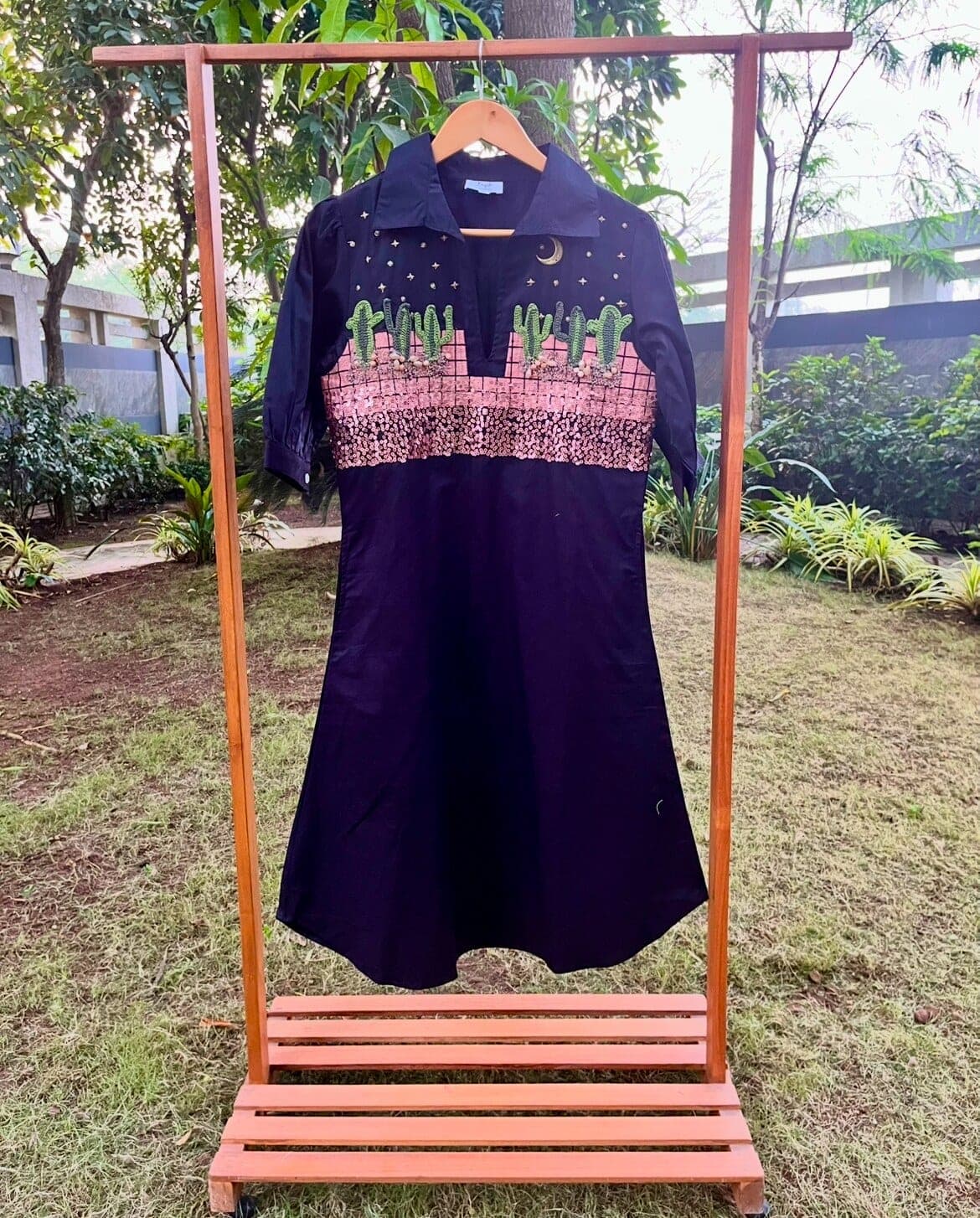 On a wooden garden rail the dress hangs on its hanger: crescent moon at the placket, cacti and rose-gold sequins across the yoke.
