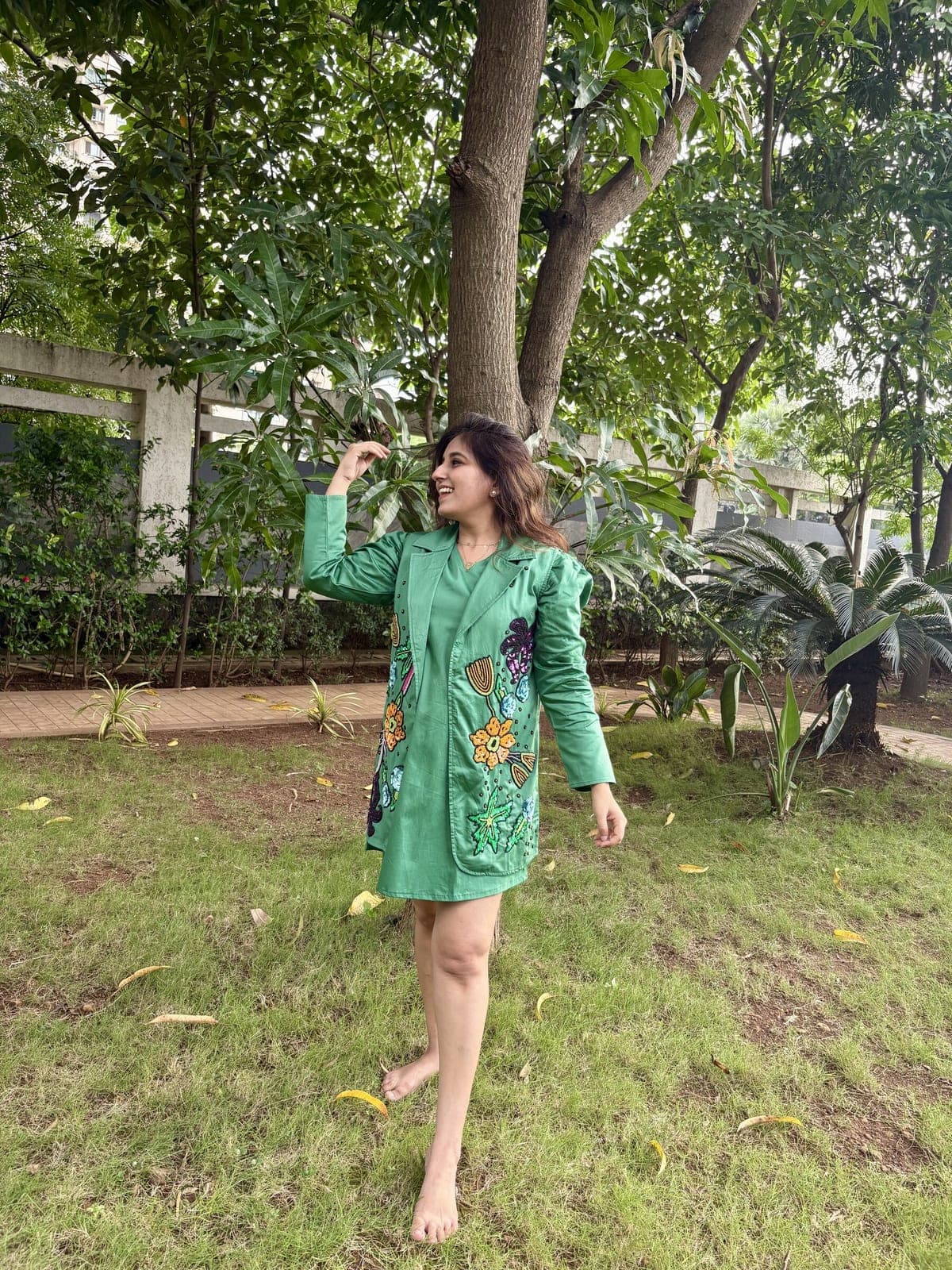 Model in a garden wearing the emerald sequin jacket, orange, blue and purple flowers worked down its front, open over a plain green dress.