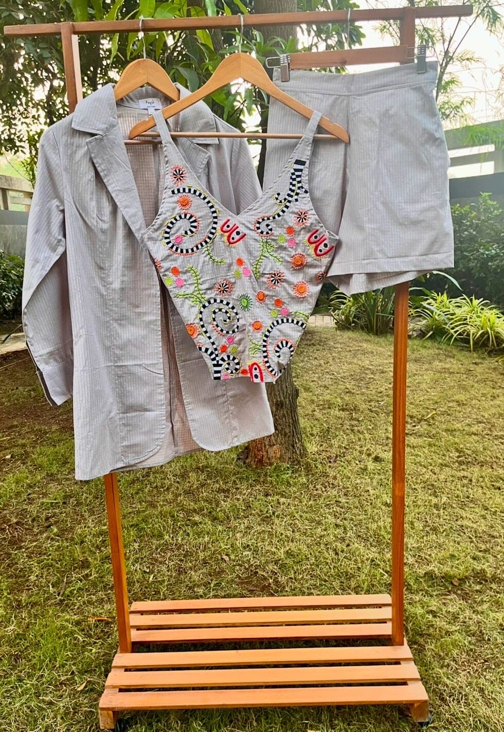 Striped grey blazer, embroidered sleeveless top with black-and-white spirals and small red faces, and matching shorts on a wooden rack.