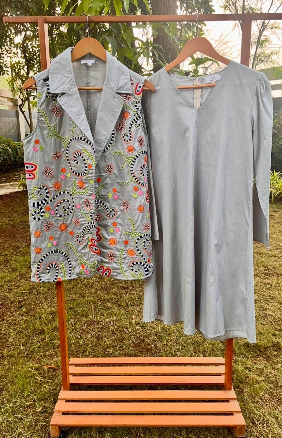 The embroidered lapel jacket and the plain grey long-sleeved dress hung side by side on a wooden rack in a garden.