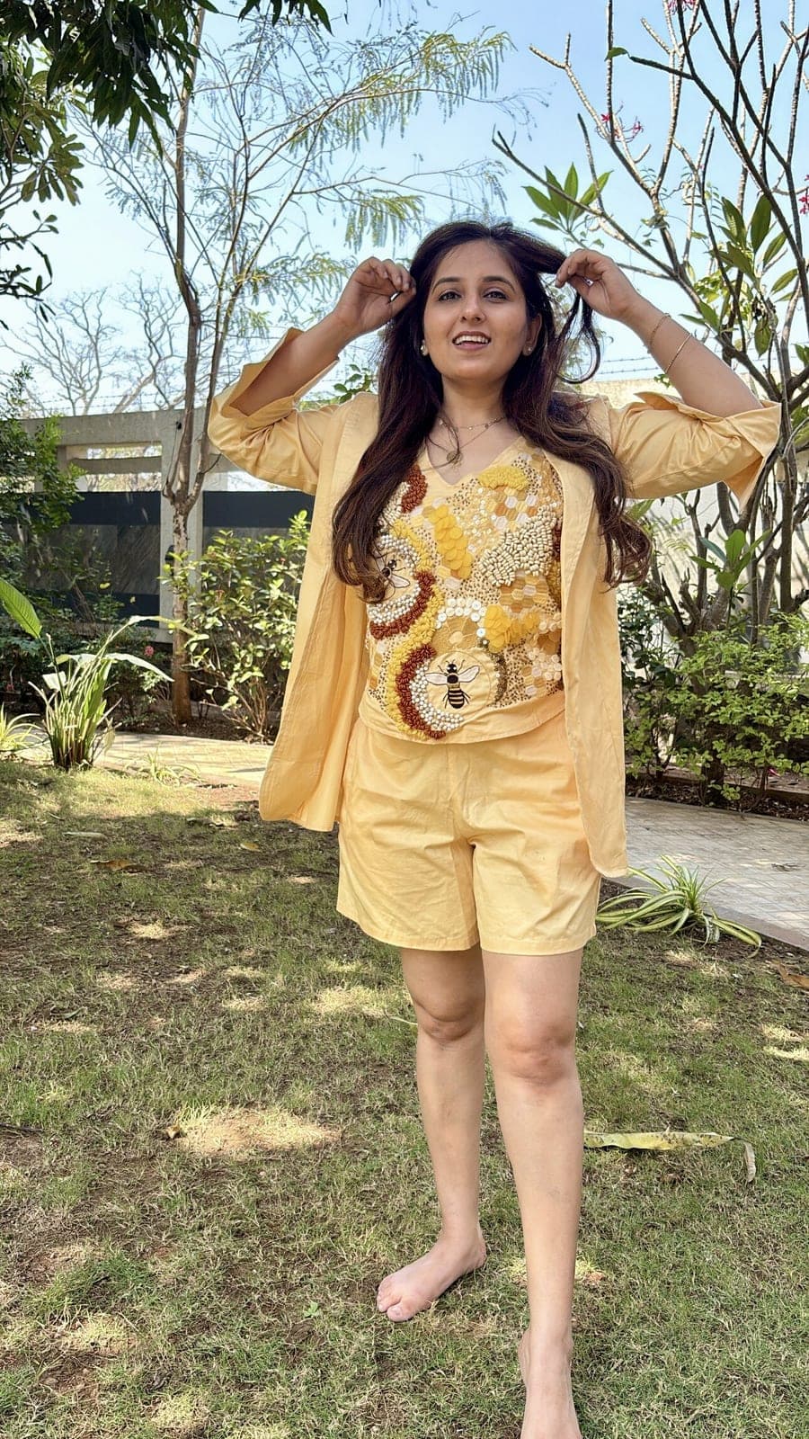 Butter-yellow blazer worn open over the bee-embroidered V-neck top and matching shorts, photographed on grass in a garden.