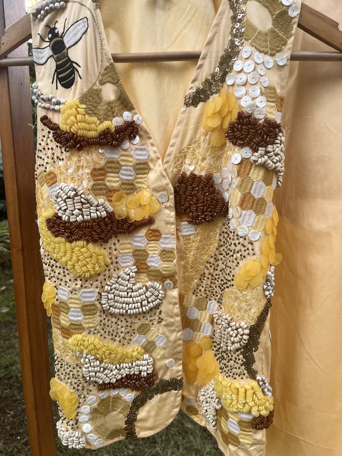 Close view of the beadwork on a wooden hanger — an embroidered bee at the shoulder above honeycomb hexagons and mother-of-pearl discs.