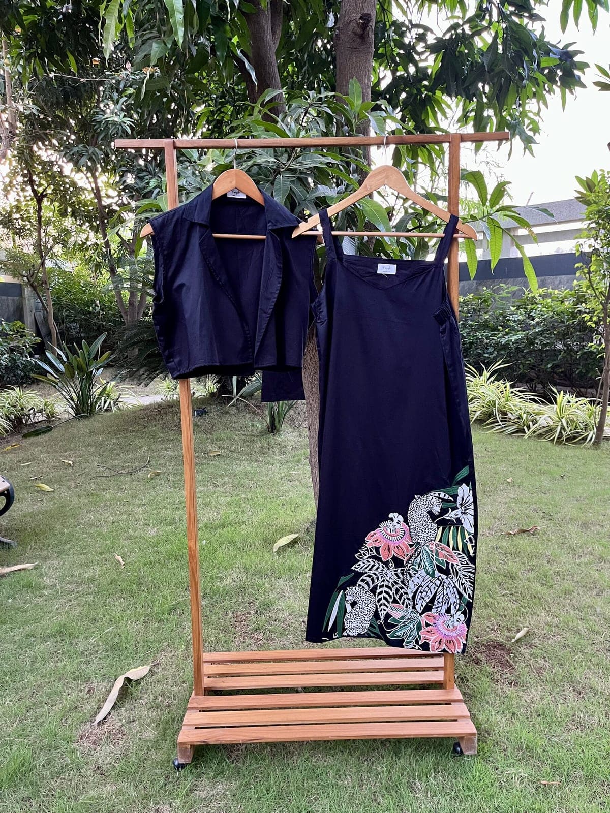 Black strappy calf-length dress hanging outdoors beside a cropped collared jacket, leopards and pink passion flowers embroidered across its lower right side.