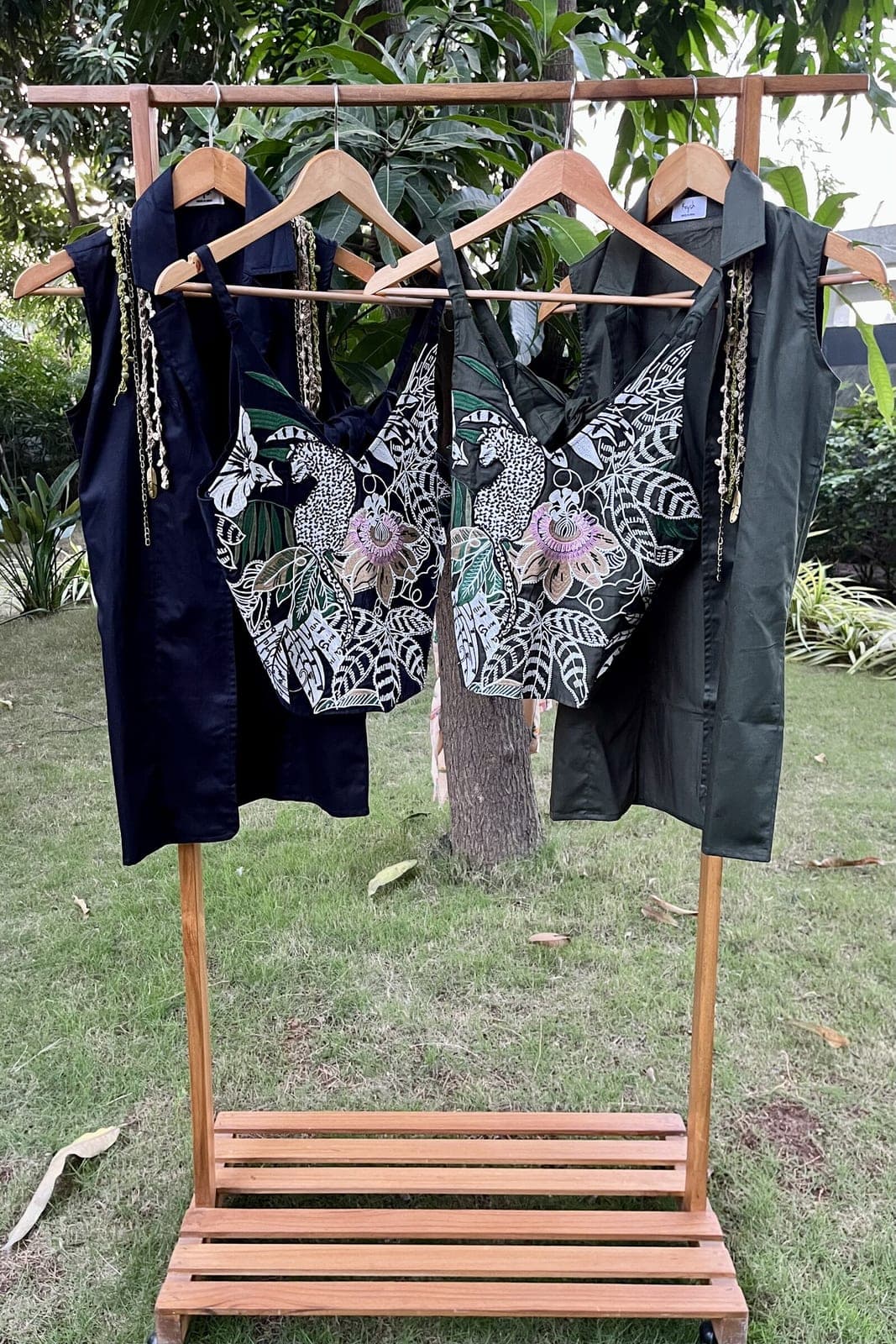 Both colourways hang together on a garden rail — black and dark green three-piece sets, embroidered strappy tops under long sleeveless blazers.