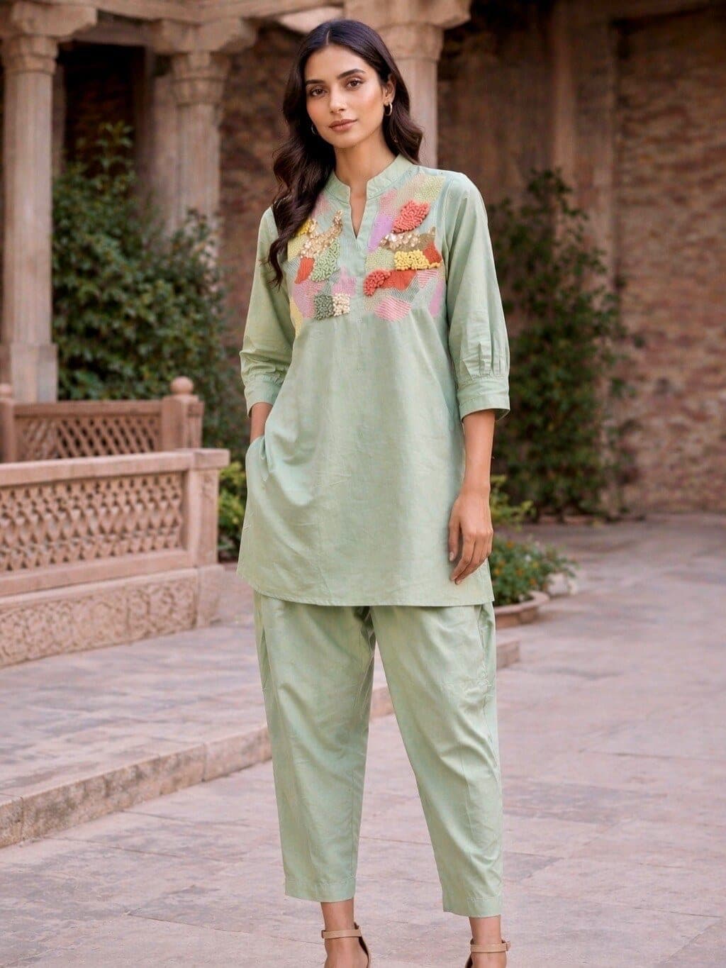 Sage green kurta set photographed in a stone courtyard, the pieced yoke in pink, coral and yellow, trousers cropped straight at the ankle.