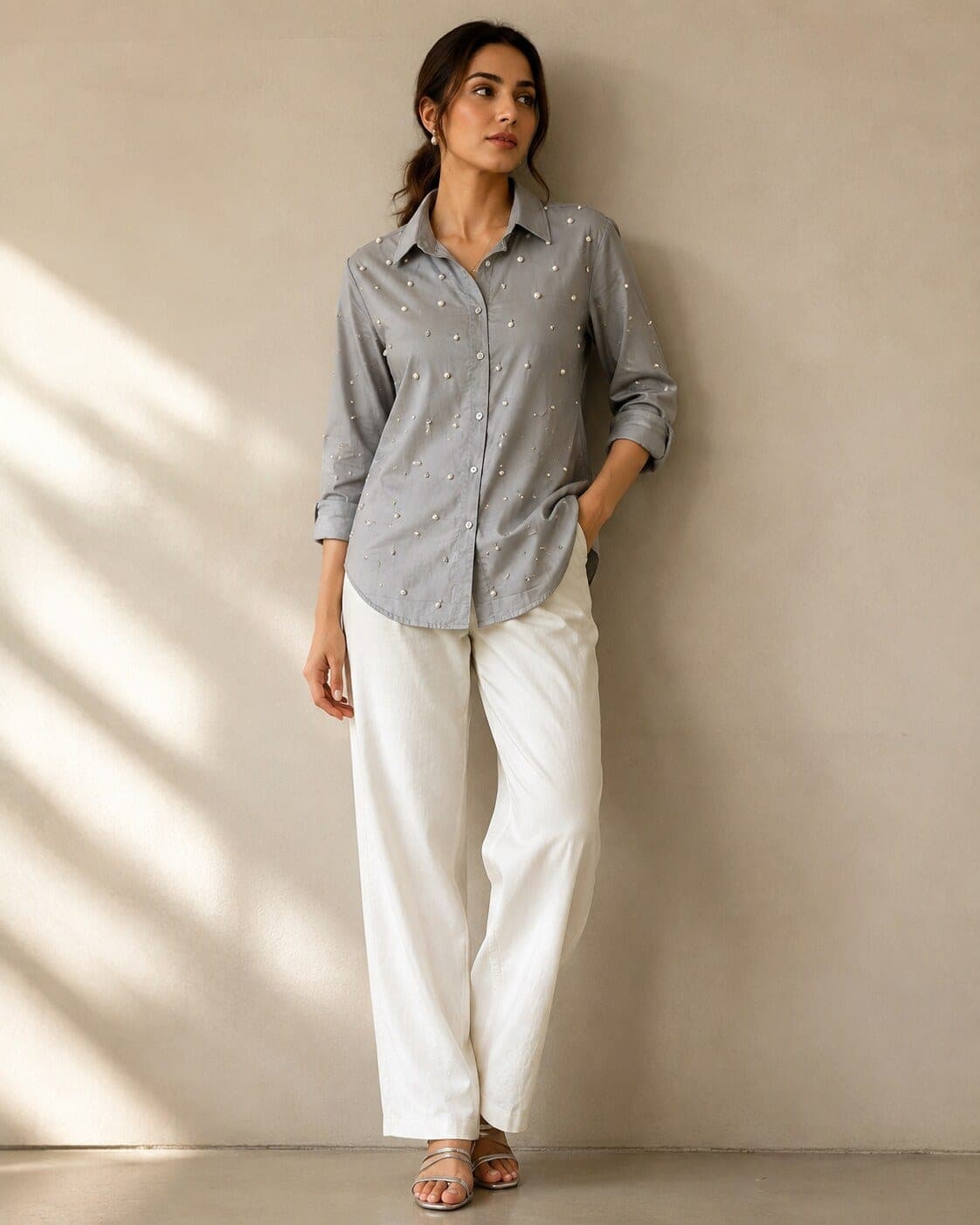 Model leans against a beige wall in the pearl-scattered grey shirt, sleeves rolled, worn loose over white wide-leg trousers.