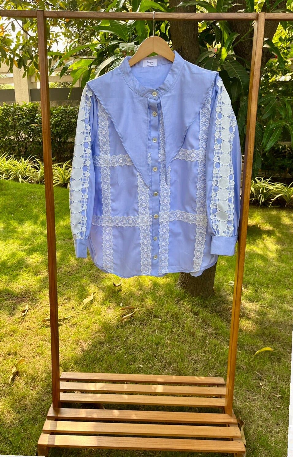 Hung on a wooden rack outdoors, a powder blue cotton shirt shows white lace panelling across the front and cutwork inserts along both sleeves.