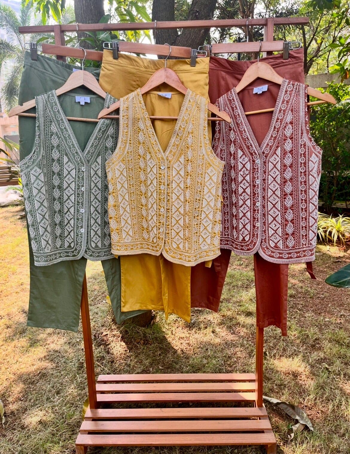 Three cord-embroidered cotton vests in olive, mustard and rust brown hang on a garden rack above their matching plain pants.