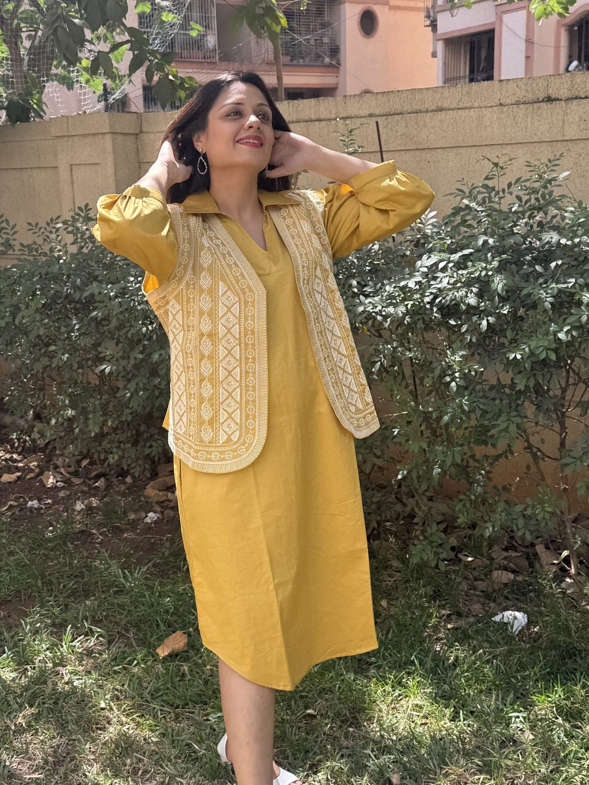 Mustard cotton A-line dress worn under an open matching vest of cream cord embroidery, three-quarter sleeves, garden hedge behind.
