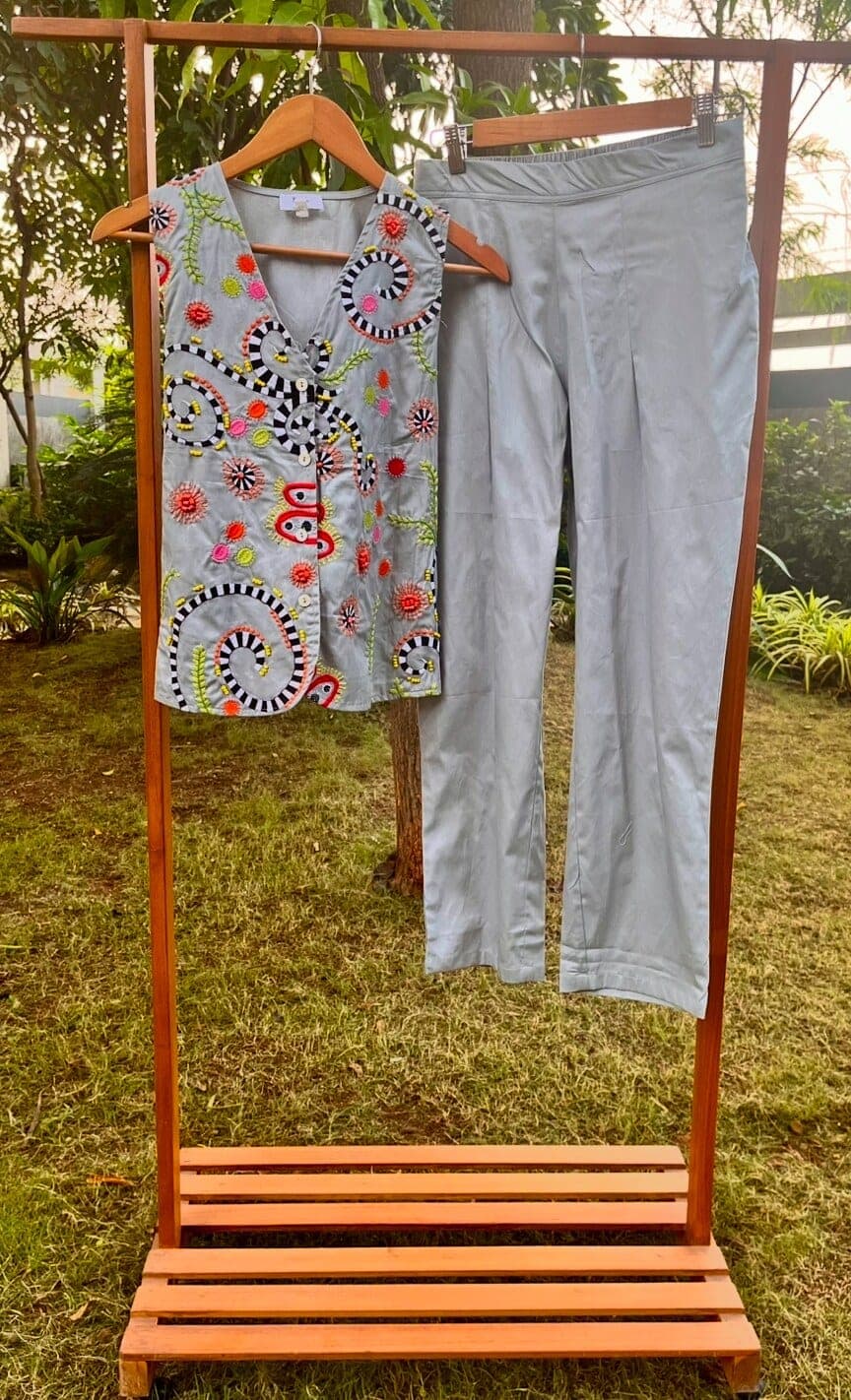 Sleeveless grey buttoned top covered in striped spirals and bright rosettes, hanging beside its matching straight-leg trousers on a wooden rack.