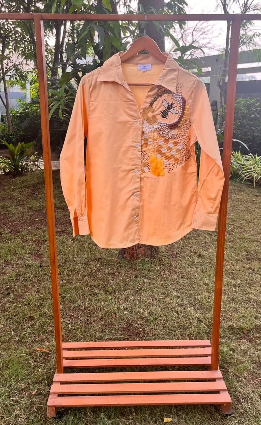 Peach cotton shirt on an outdoor rail, a beaded bee and gold honeycomb worked down its right front, sleeves and collar left plain.