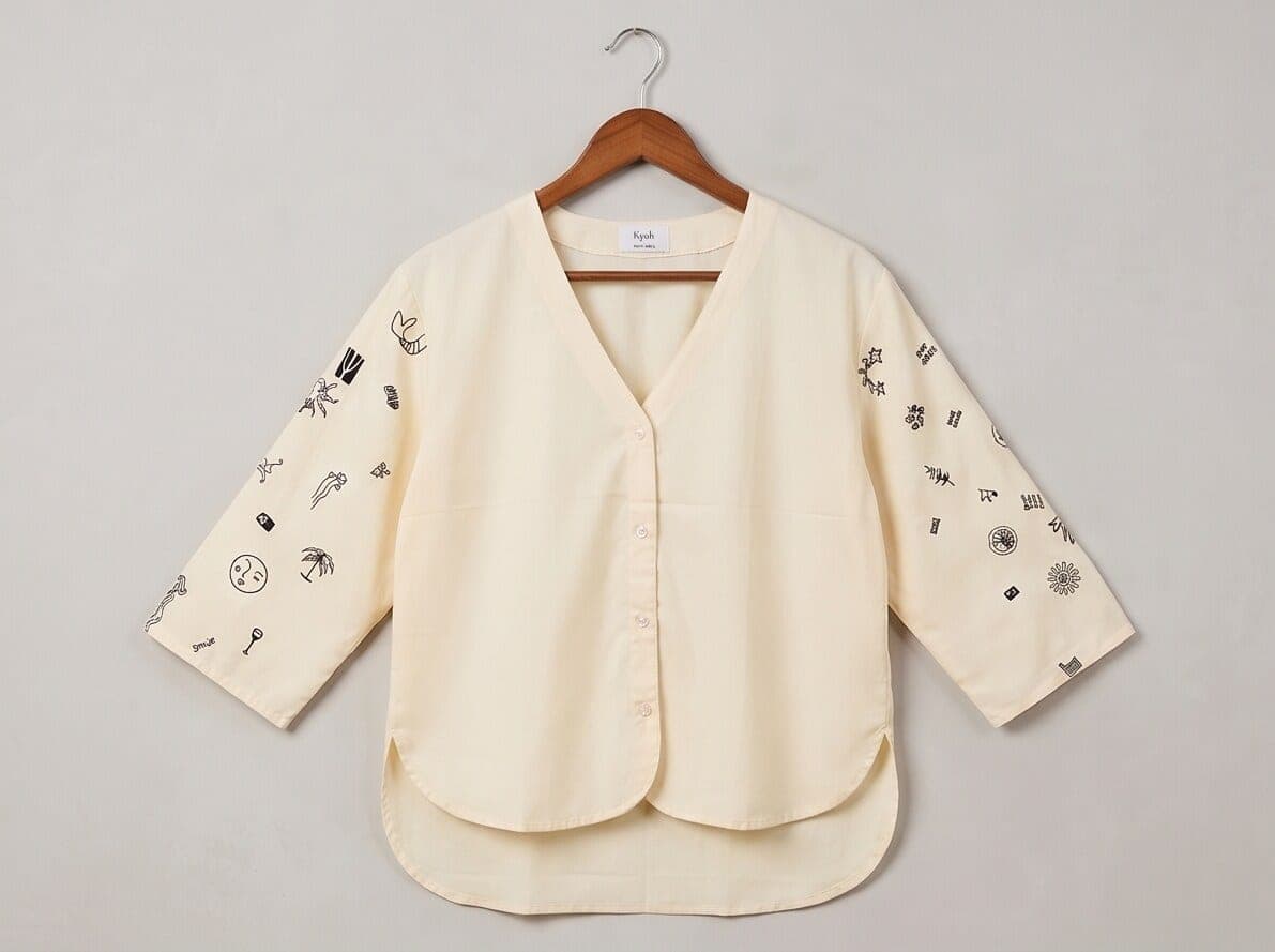 Butter yellow cotton blouse on a wooden hanger, navy icons stitched along both three-quarter sleeves, plain V-neck body and curved hem.
