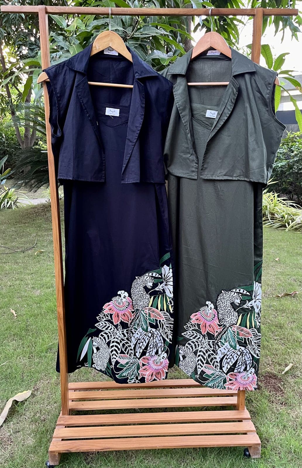 Two calf-length cotton dresses on a garden rail, black and dark green, each under a cropped sleeveless collared jacket, embroidery at the hem.