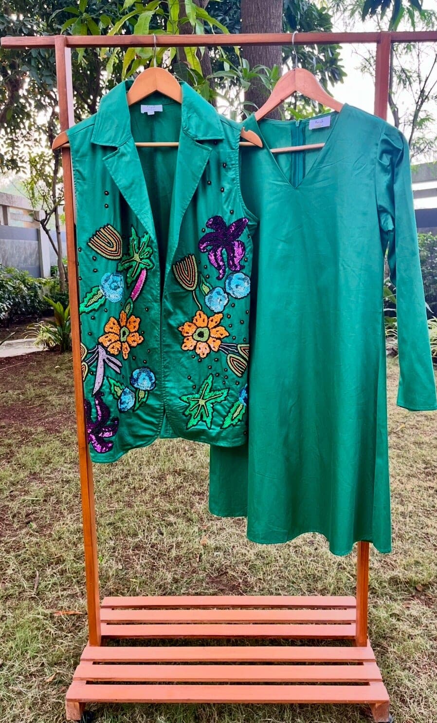 Plain emerald V-neck dress with long sleeves, hung on a wooden garden rack beside the sequin-embroidered jacket from the same collection.