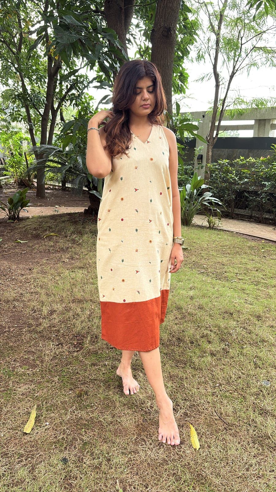 Beige cotton linen A-line dress worn barefoot in a garden, scattered floral embroidery above a rust colour-blocked hem.