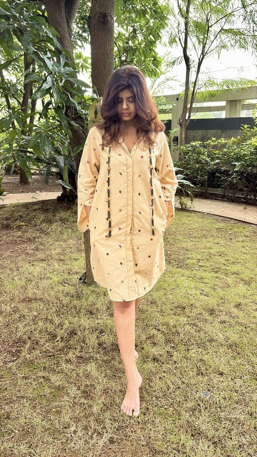 Sand-coloured cotton linen shirt dress worn in a garden, small multicoloured embroidery over the body and sleeves rolled to the elbow.