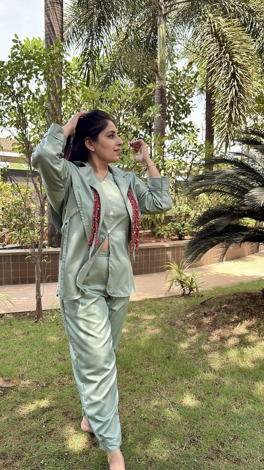Model in a garden wearing a sage viscose-blend blazer open over a matching shirt and trousers, red beaded tassels hanging from both lapels.