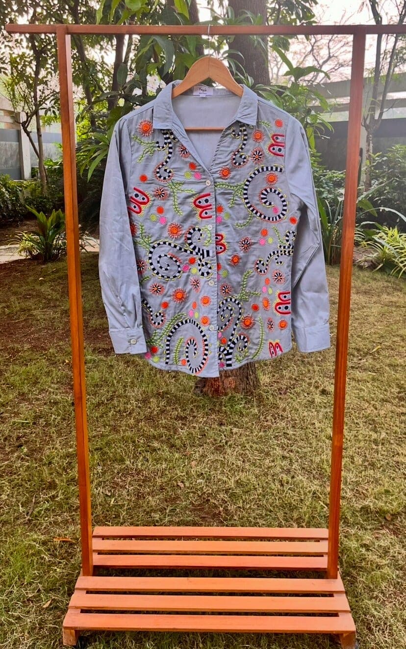 Light grey shirt on an outdoor wooden rack, front covered in striped coils, orange starbursts and small embroidered beetles.