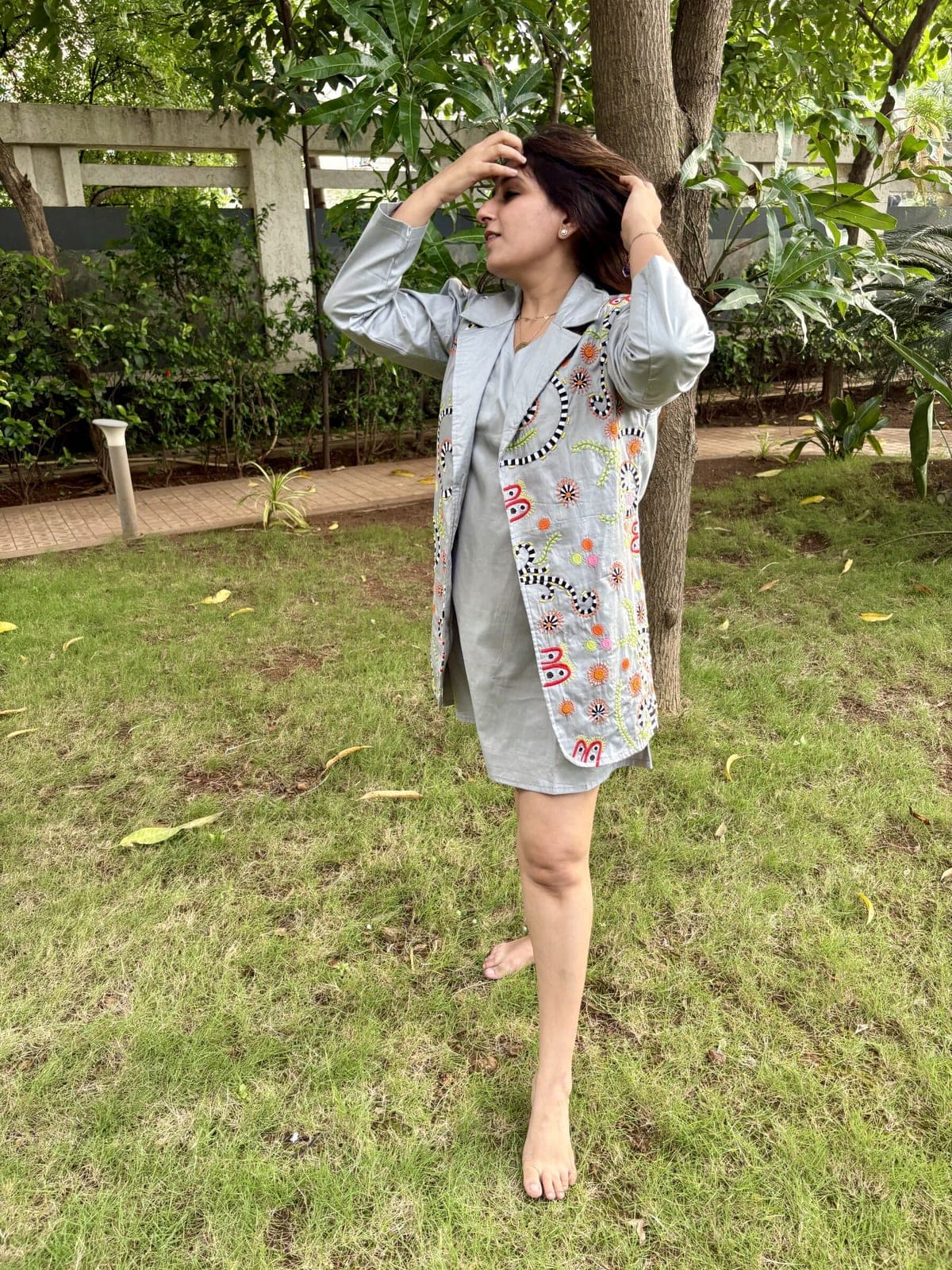 Model on grass in a plain grey knee-length shift dress under a sleeveless grey jacket embroidered with striped coils and orange sunbursts.